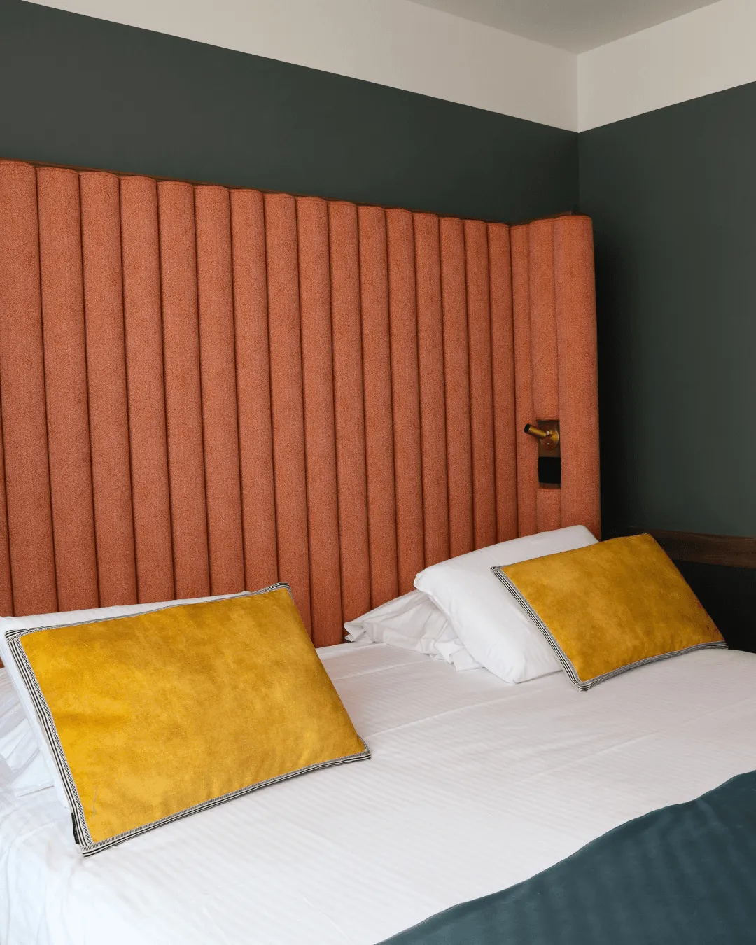 Bett mit orangefarbenem Kopfteil, zwei gelben Kissen und weißen Laken in einem Zimmer mit dunklen Wänden