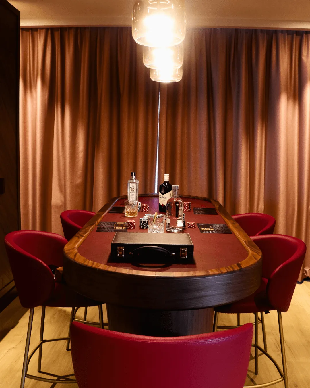 Pokertisch aus Holz mit sechs roten Stühlen, dekoriert mit Flaschen, Gläsern und Chips, beleuchtet von einer Hängeleuchte in einem Raum mit goldenen Vorhängen