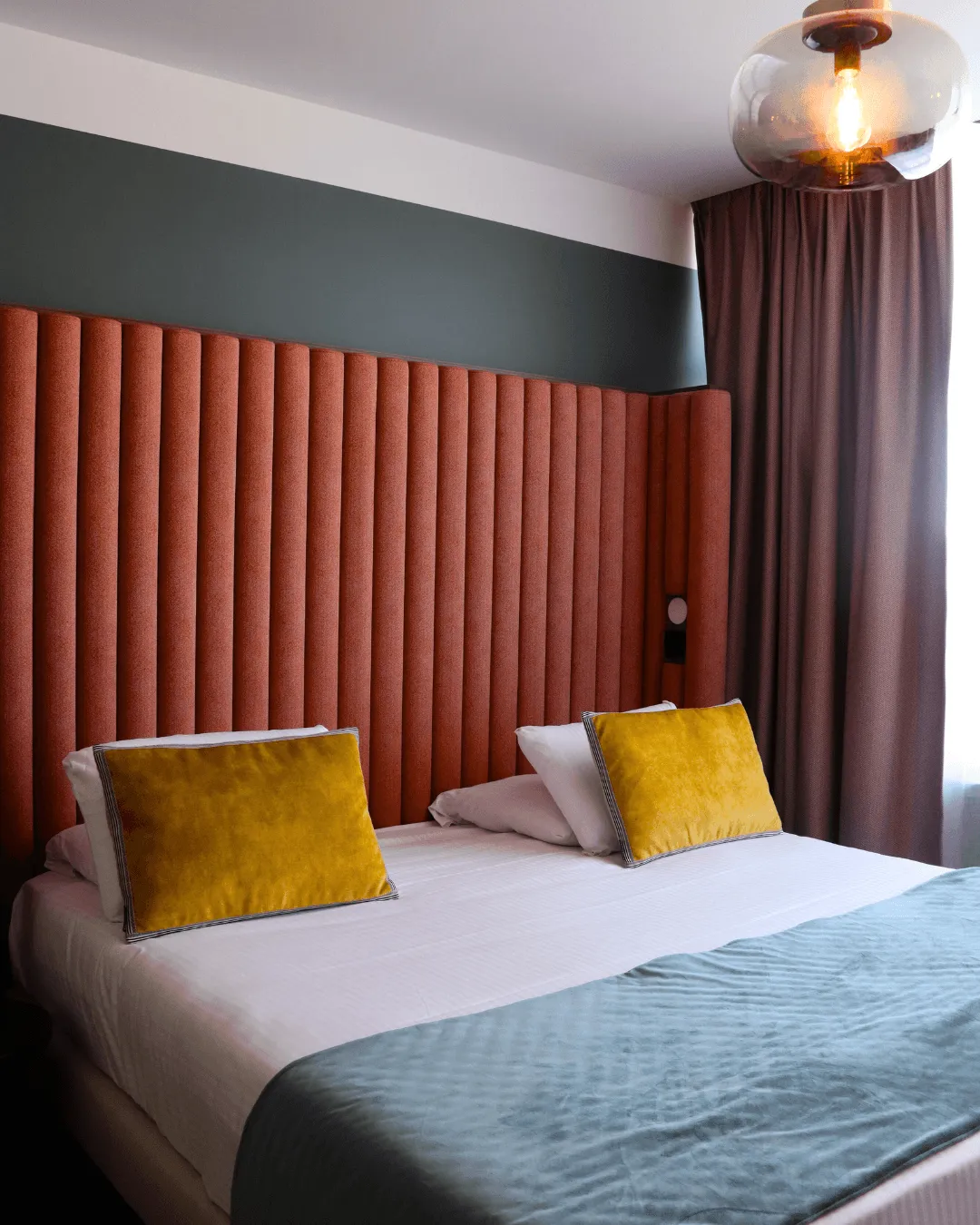 Bett mit rot gestreiftem Kopfteil, weißen Laken und grauer Decke, zwei gelben Kissen und einer eingeschalteten Kugellampe an der Decke