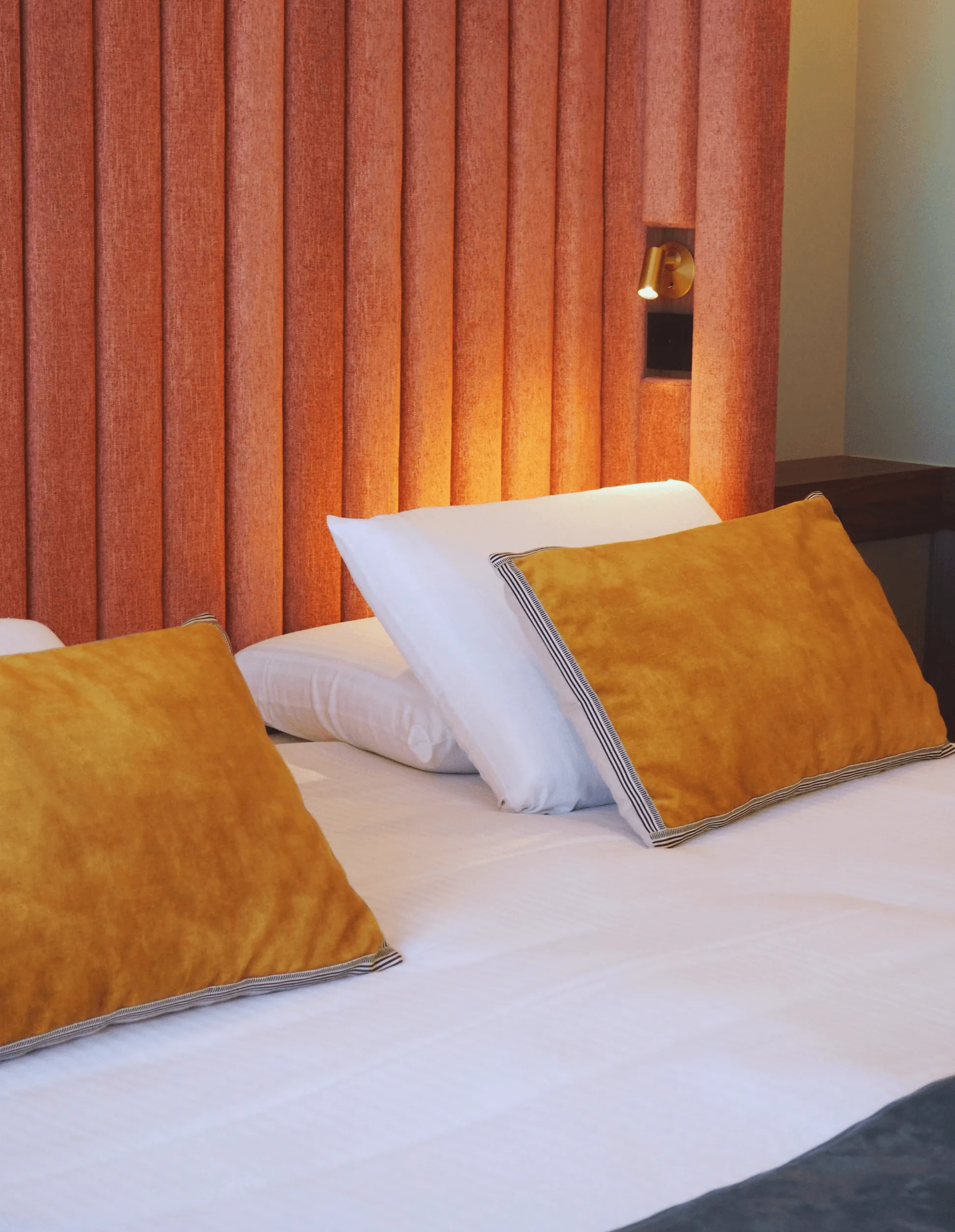 Bett mit weißen Laken und orange-weißen Kissen, eine Wand mit orangefarbenen Vorhängen und eine eingeschaltete Nachttischlampe