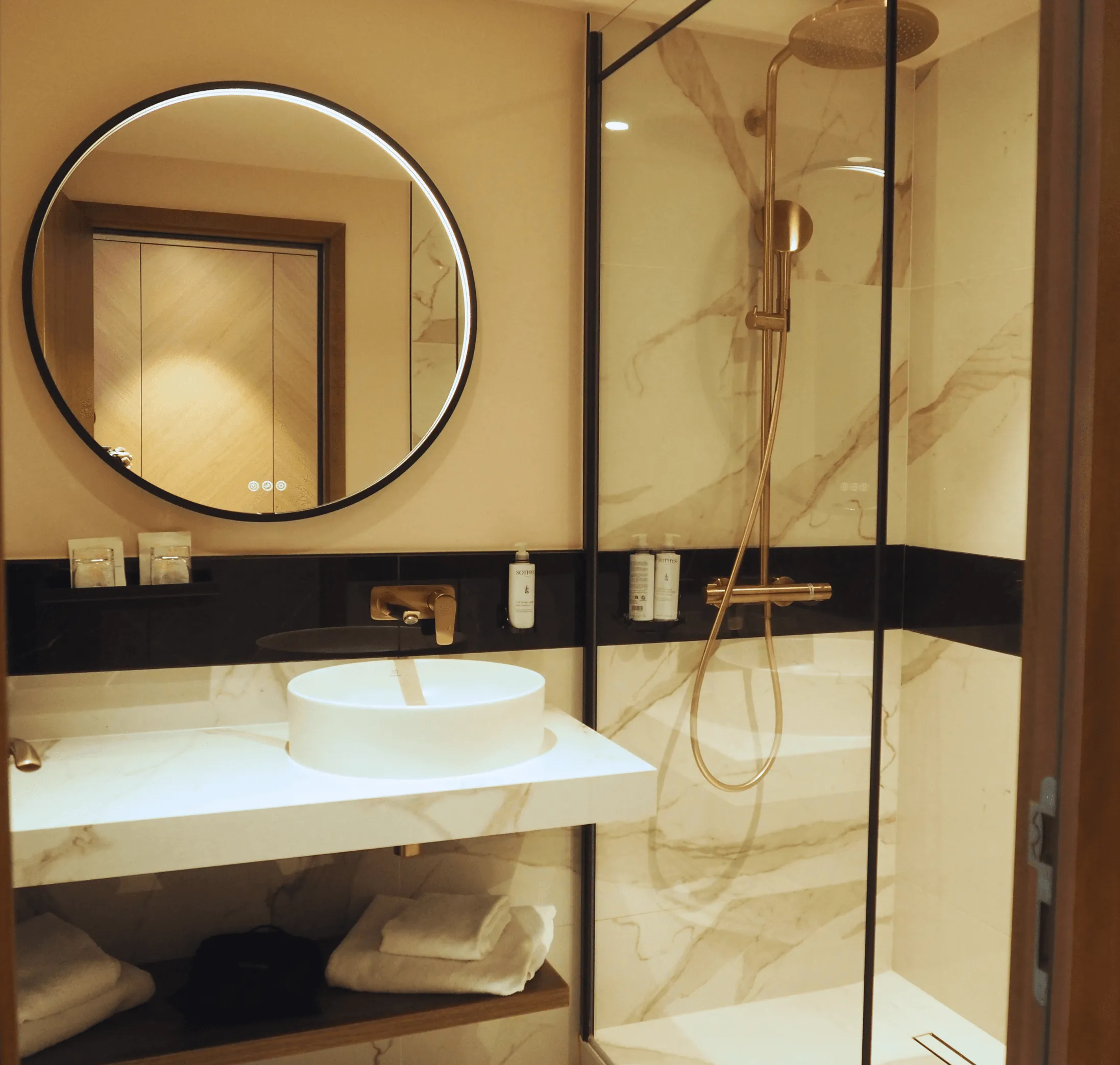 Badezimmer mit weißem Waschbecken, beleuchtetem Rundspiegel und Duschkabine mit Glaswand