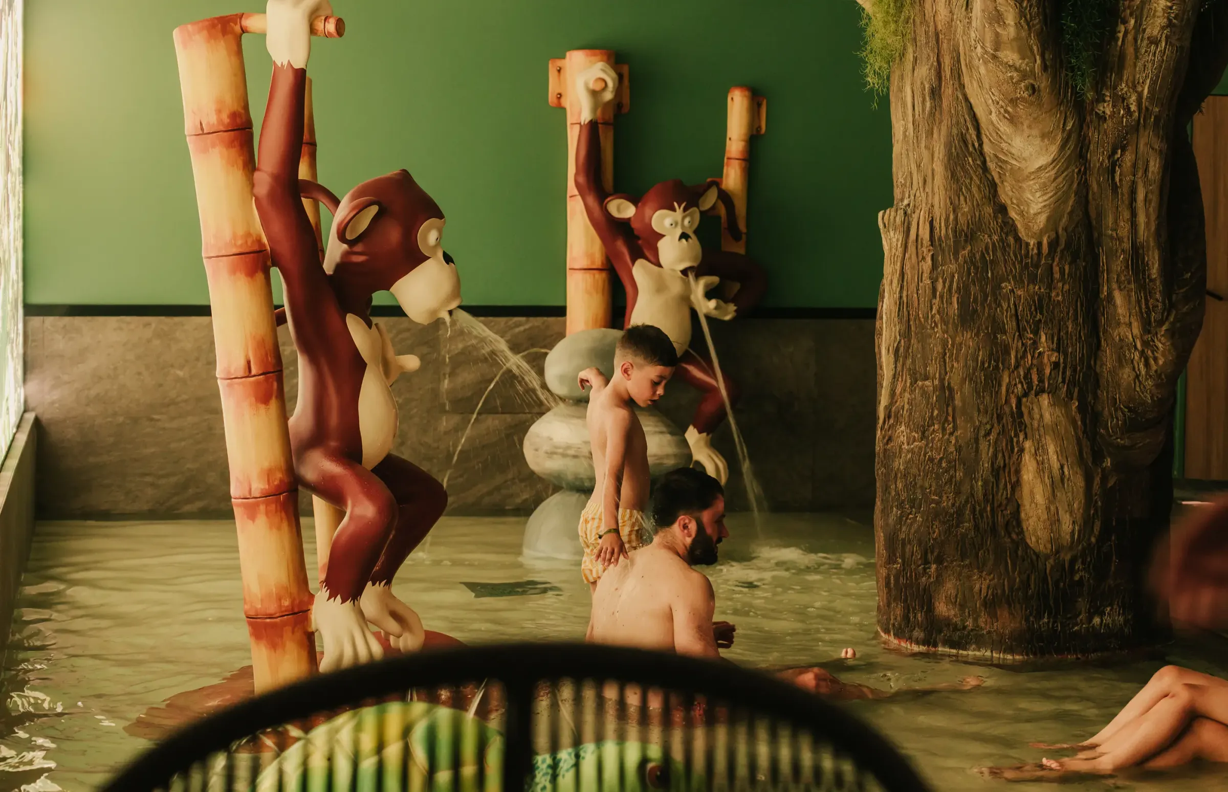 Des personnes se détendent dans un bain thermal décoré avec des sculptures de singes et des bambous, avec des jets d'eau en arrière-plan