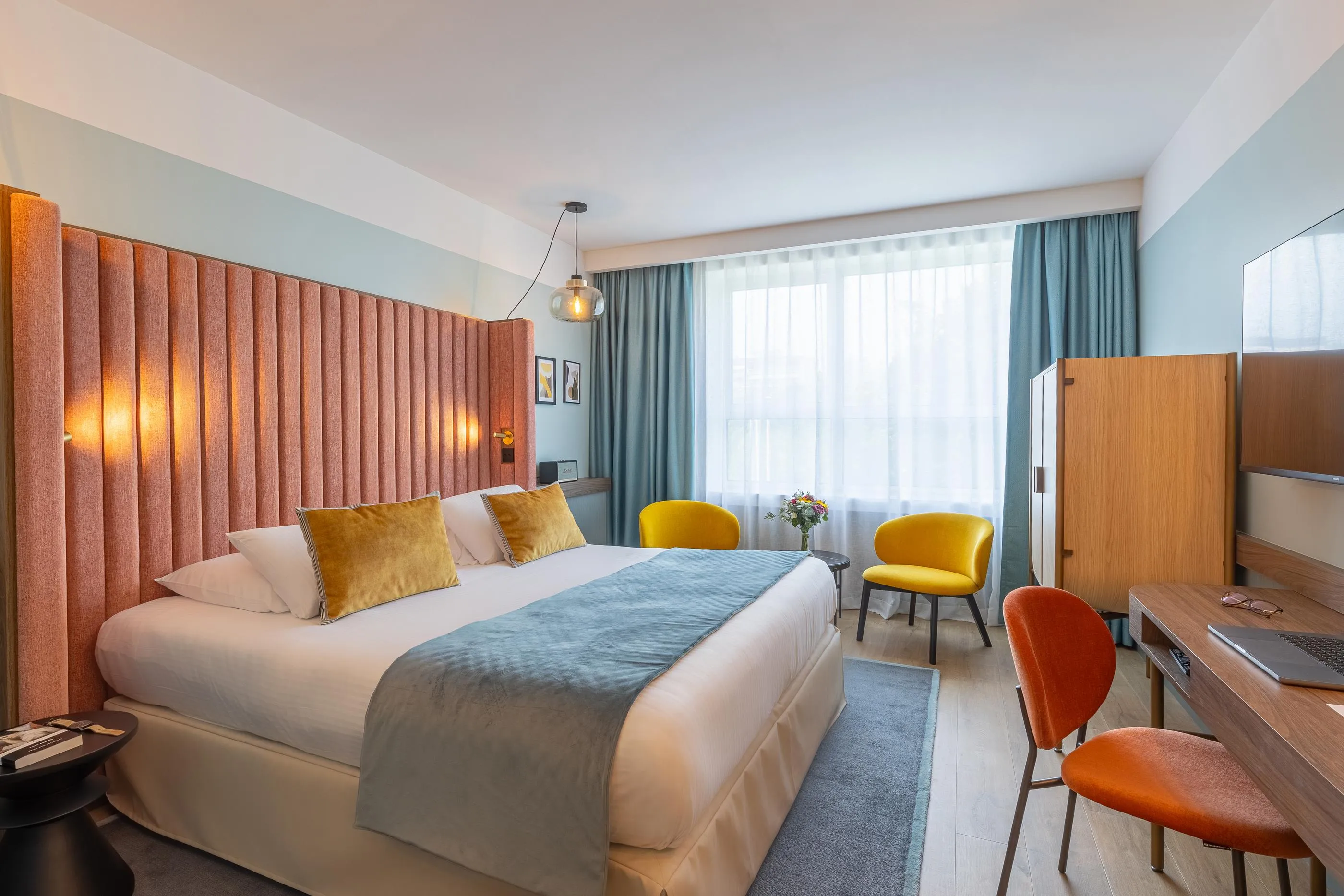 Hotelzimmer mit einem Doppelbett, zwei bunten Stühlen, einem Schreibtisch und großen Fenstern mit Vorhängen