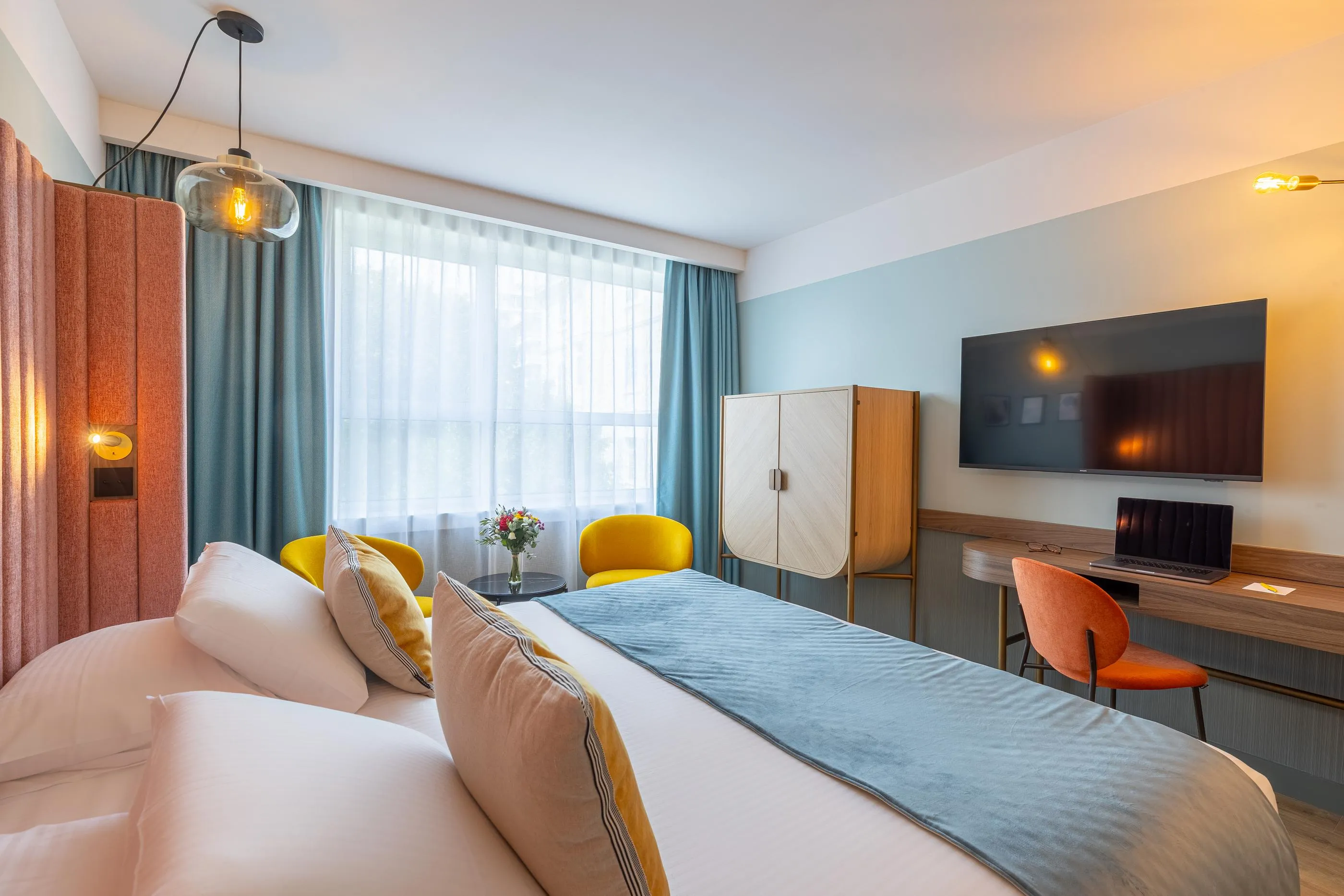 Hotelzimmer mit einem Doppelbett, einem Fernseher, einem Schreibtisch und einem großen Fenster mit Vorhängen