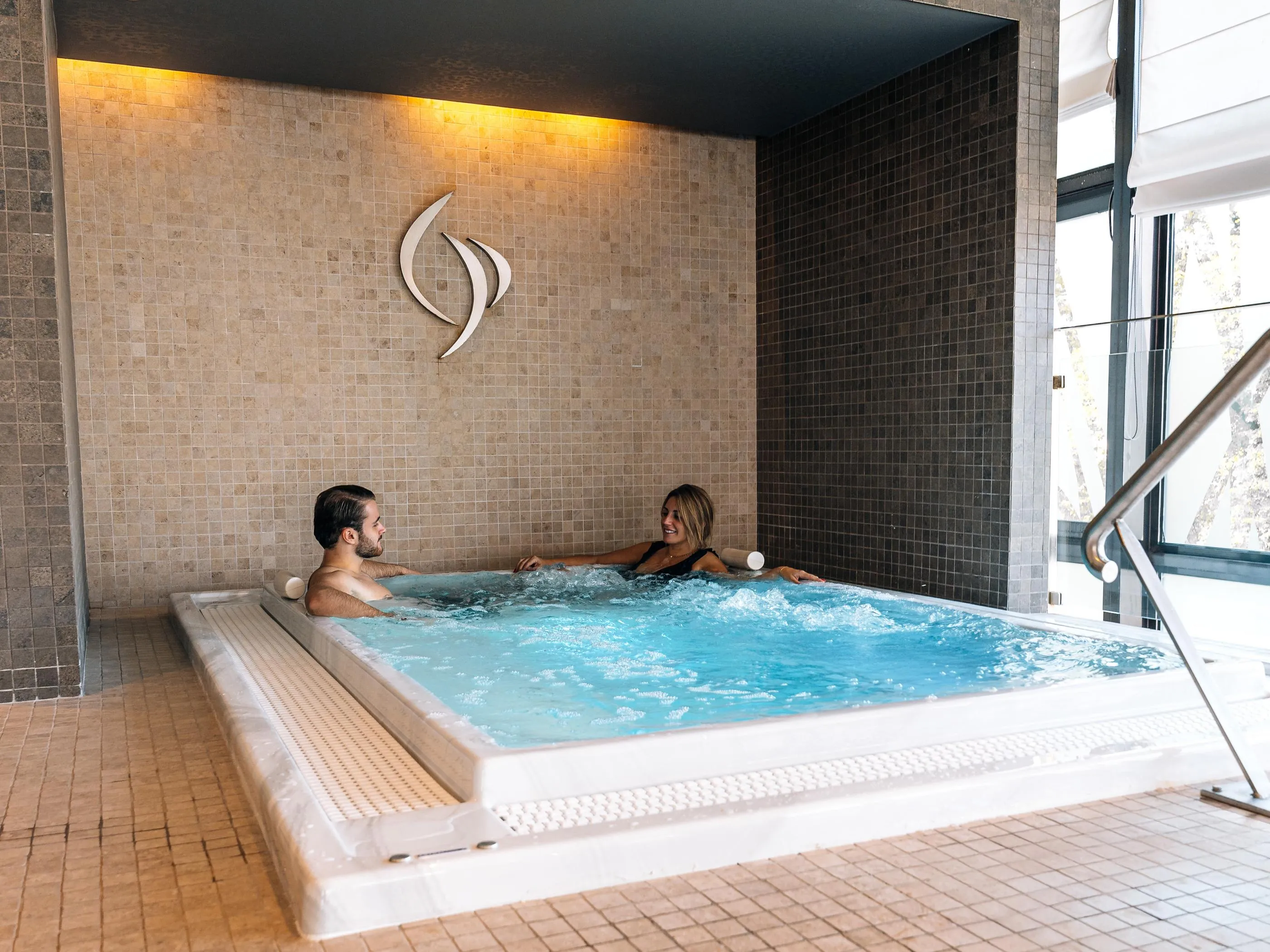 Zwei Personen sitzen in einem Whirlpool, umgeben von Fliesen, mit einem großen Fenster in der Nähe.