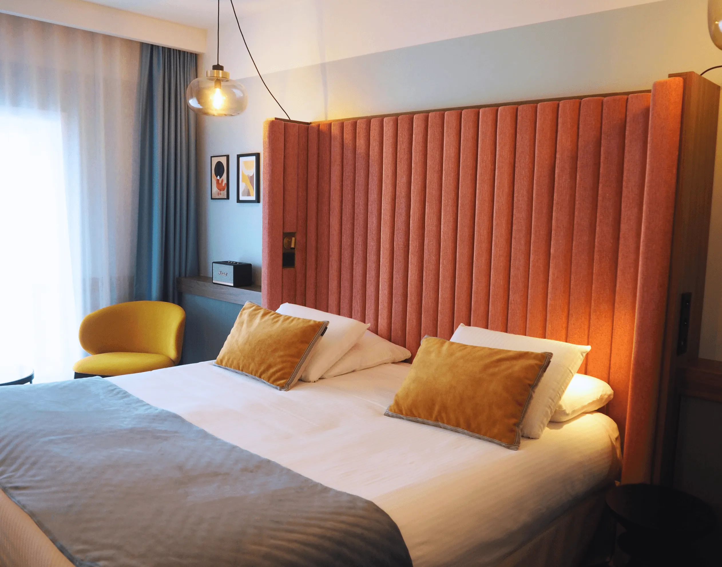 Zimmer mit einem Doppelbett mit weißen Laken und orangen Kissen, einem roten Samtkopfteil, einem gelben Stuhl und einer Hängelampe